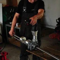 Pracownik w kombinezonie roboczym montuje element konstrukcyjny studni abisyńskiej przy użyciu specjalistycznego narzędzia elektrycznego w warsztacie.