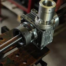 Szczegółowe ujęcie przekładni ślimakowej z wałem na drewnianym stole warsztatowym, widoczne śruby mocujące i aluminiowa obudowa.