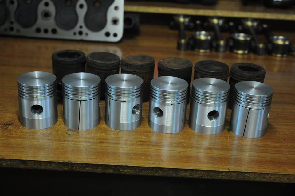 Rząd nowych, srebrnych tłoków silnikowych z widocznymi otworami i rowkami, ustawionych obok rzędu starych, zużytych tłoków, na tle głowicy silnika i innych części.