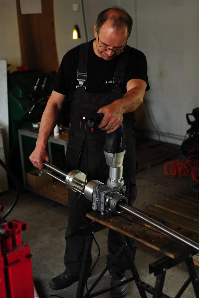 Pracownik w kombinezonie roboczym montuje element konstrukcyjny studni abisyńskiej przy użyciu specjalistycznego narzędzia elektrycznego w warsztacie.