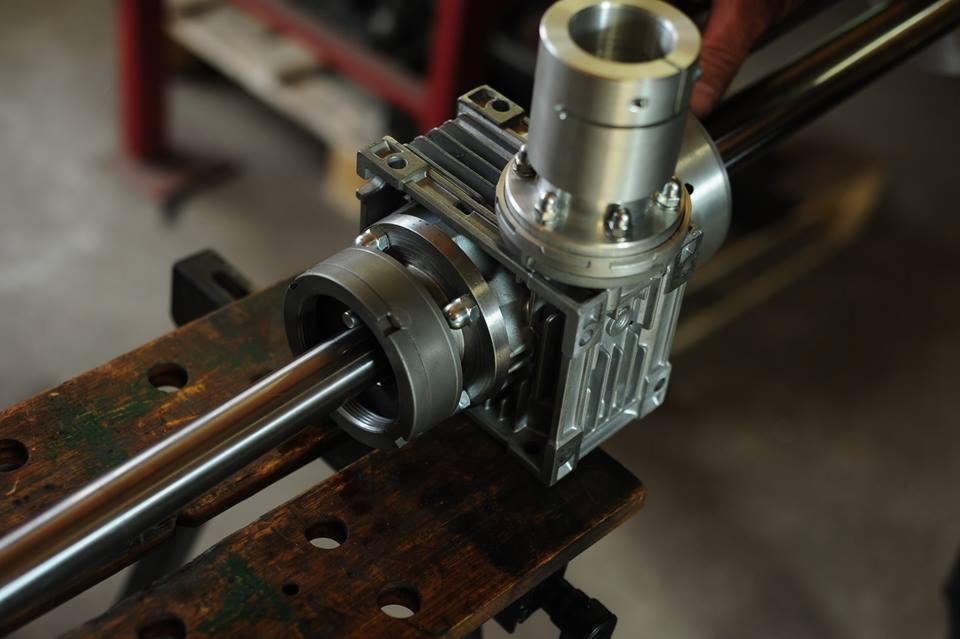 Szczegółowe ujęcie przekładni ślimakowej z wałem na drewnianym stole warsztatowym, widoczne śruby mocujące i aluminiowa obudowa.