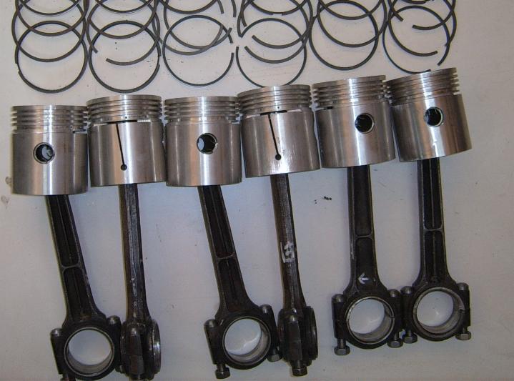 Sześć tłoków silnikowych z korbowodami ułożonych obok siebie, z pierścieniami tłokowymi w tle, na jasnym tle.
