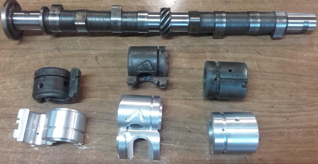 Wałek rozrządu i elementy metalowe, prawdopodobnie części do wiertnicy, leżące na drewnianej powierzchni.