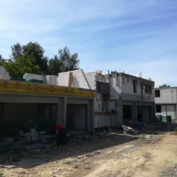 Wznoszenie osiedla domów jednorodzinnych z betonu komórkowego, widoczne zbrojenia i szalunki, teren budowy z gruzem i narzędziami, pochmurne niebo.