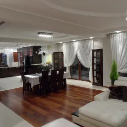 Przestronne wnętrze domu z otwartą kuchnią, jadalnią i salonem; parkiet z ciemnego drewna, białe ściany i zasłony, kryształowe żyrandole, meble w odcieniach brązu i bieli.