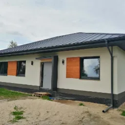 Parterowy dom z szarą blachodachówką, jasną elewacją i drewnianymi okiennicami, widoczne elementy wykończeniowe i teren wokół domu w trakcie zagospodarowywania.