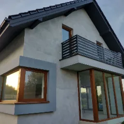 Nowoczesny dom z oknami PCV w kolorze drewna, balkon z czarną, poziomą balustradą i przeszklonym tarasem, widok z zewnątrz o zmierzchu.