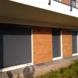 Nowoczesny budynek jednorodzinny z zamontowanymi szarymi roletami zewnętrznymi i oknami, wykończony drewnopodobną elewacją i elementami z jasnego tynku.