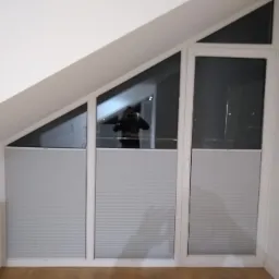 Trzyczęściowe okno PCV na poddaszu z zamontowanymi plisowanymi roletami wewnętrznymi w kolorze jasnoszarym, częściowo zasłoniętymi, z widocznym odbiciem fotografa w szybie.