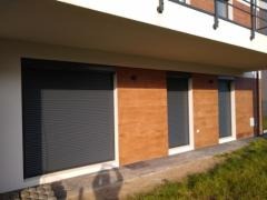 Nowoczesny budynek jednorodzinny z zamontowanymi szarymi roletami zewnętrznymi i oknami, wykończony drewnopodobną elewacją i elementami z jasnego tynku.