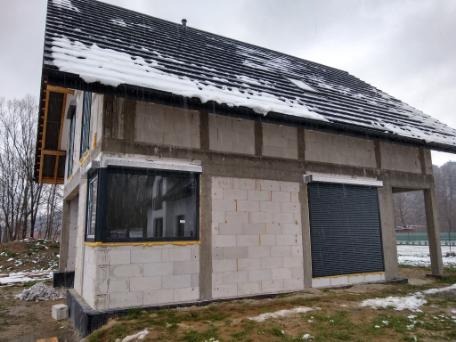 Surowy stan budynku w trakcie budowy z zamontowanymi oknami z czarną ramą i zewnętrzną roletą w kolorze grafitowym, widok z zewnątrz w pochmurny dzień.