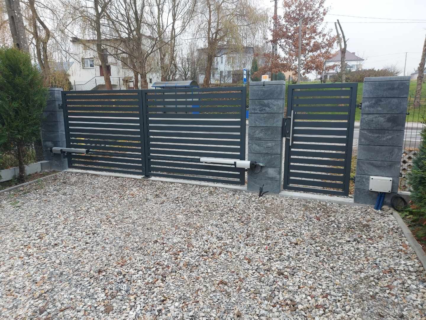 Nowoczesna, pozioma brama wjazdowa z automatyką Somfy, zamontowana na posesji z wysypanym żwirem podjazdem i betonowymi słupkami.