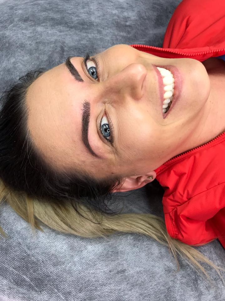 Kobieta z nowymi, ciemnymi brwiami po zabiegu makijażu permanentnego; widoczne jasne włosy u nasady i czerwona kurtka.