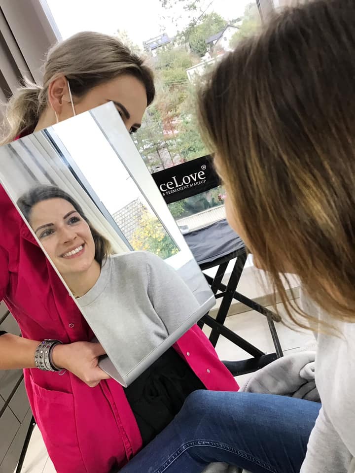 Klientka uśmiecha się, patrząc w lustro trzymane przez kosmetyczkę w różowym uniformie, w tle widoczny budynek przez okno.