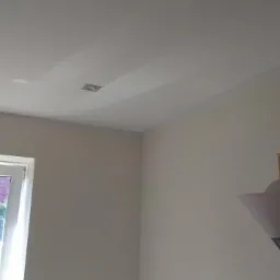 Świeżo pomalowane wnętrze z delikatnym odcieniem szarości na ścianach i suficie, widoczne okno z widokiem na zieleń oraz lampa podłogowa z abażurem w kształcie kielicha.