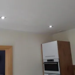 Wykończona kuchnia z białymi szafkami i drewnianymi akcentami, widoczne dwa kwadratowe reflektory w suficie i okap nad płytą grzewczą.