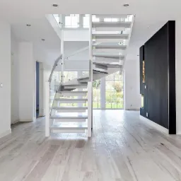 Nowoczesne, spiralne schody drewniane z białymi stopniami i szklaną balustradą w jasnym, minimalistycznym wnętrzu z parkietem o naturalnym odcieniu drewna.