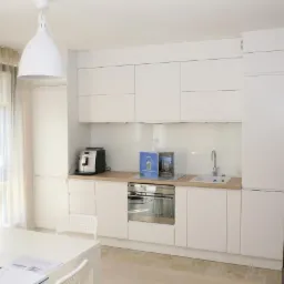 Jasna, minimalistyczna kuchnia z białymi frontami, drewnianym blatem i sprzętem AGD. Widoczne dwa wiszące, białe klosze lamp. Na stole leżą dokumenty i niebieska ozdoba.