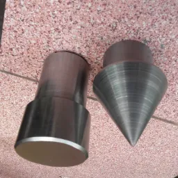 Dwa metalowe elementy: walec z kołnierzem i stożek, leżące na różowej, nakrapianej płytce.