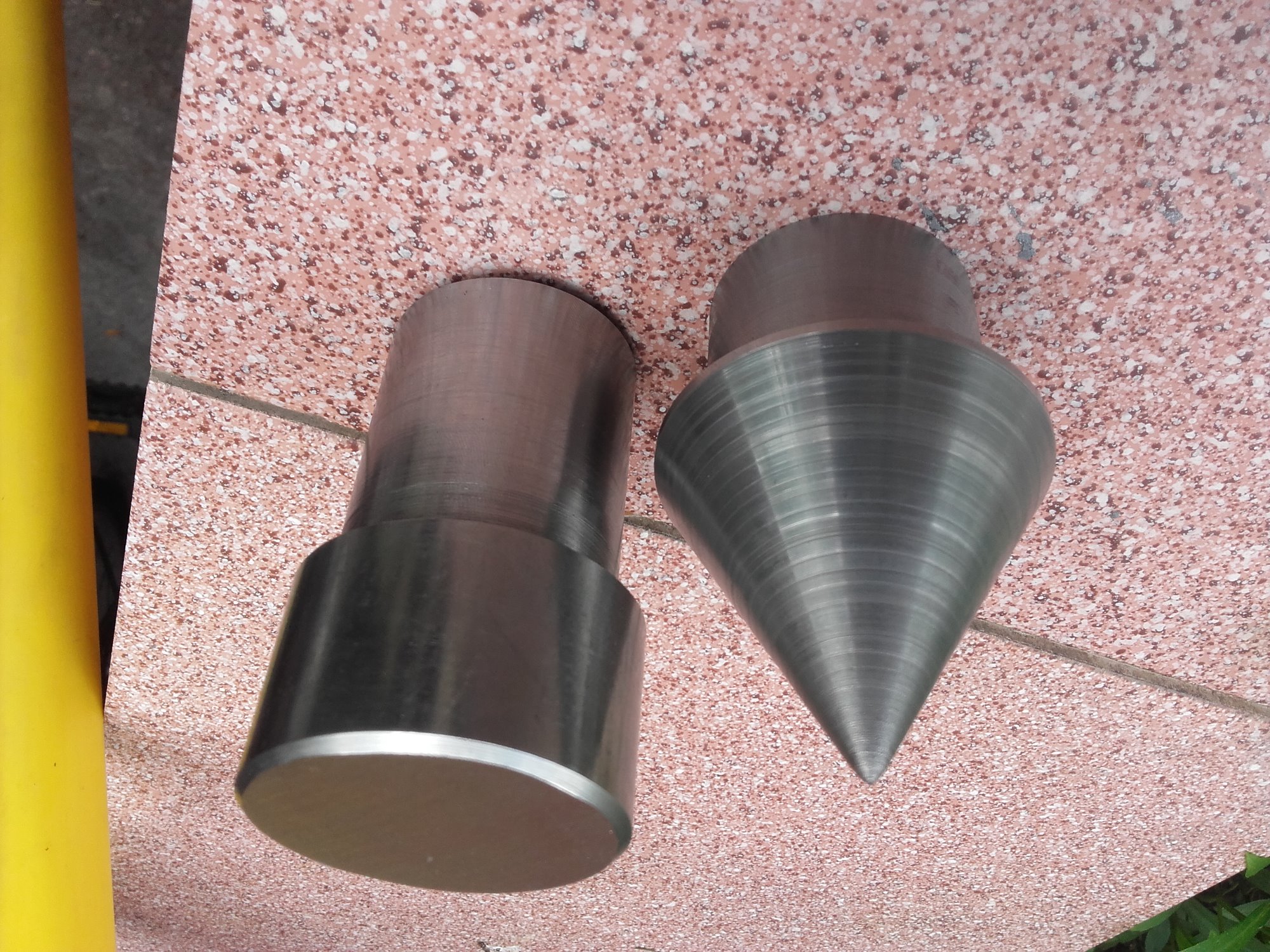 Dwa metalowe elementy: walec z kołnierzem i stożek, leżące na różowej, nakrapianej płytce.