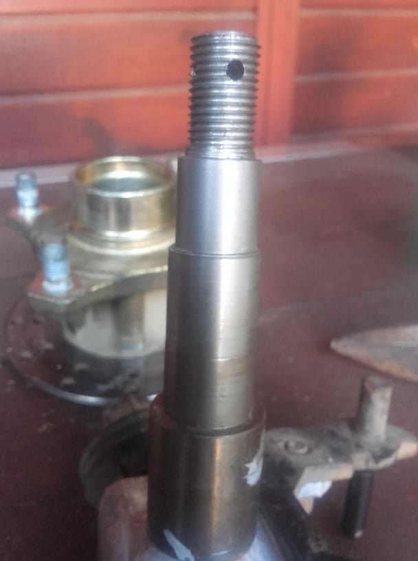 Metalowy trzpień z gwintem i otworem, obok element metalowy z otworami na śruby, na tle drewnianej ściany.