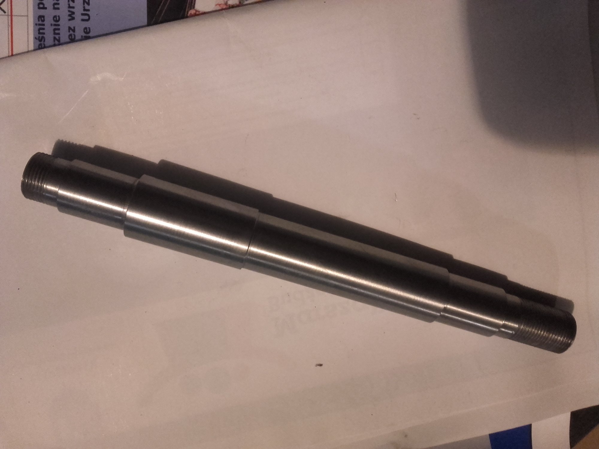 Metalowy wał z gwintowanymi końcami i stopniami o różnej średnicy, leżący na białym tle z fragmentem tekstu w rogu.