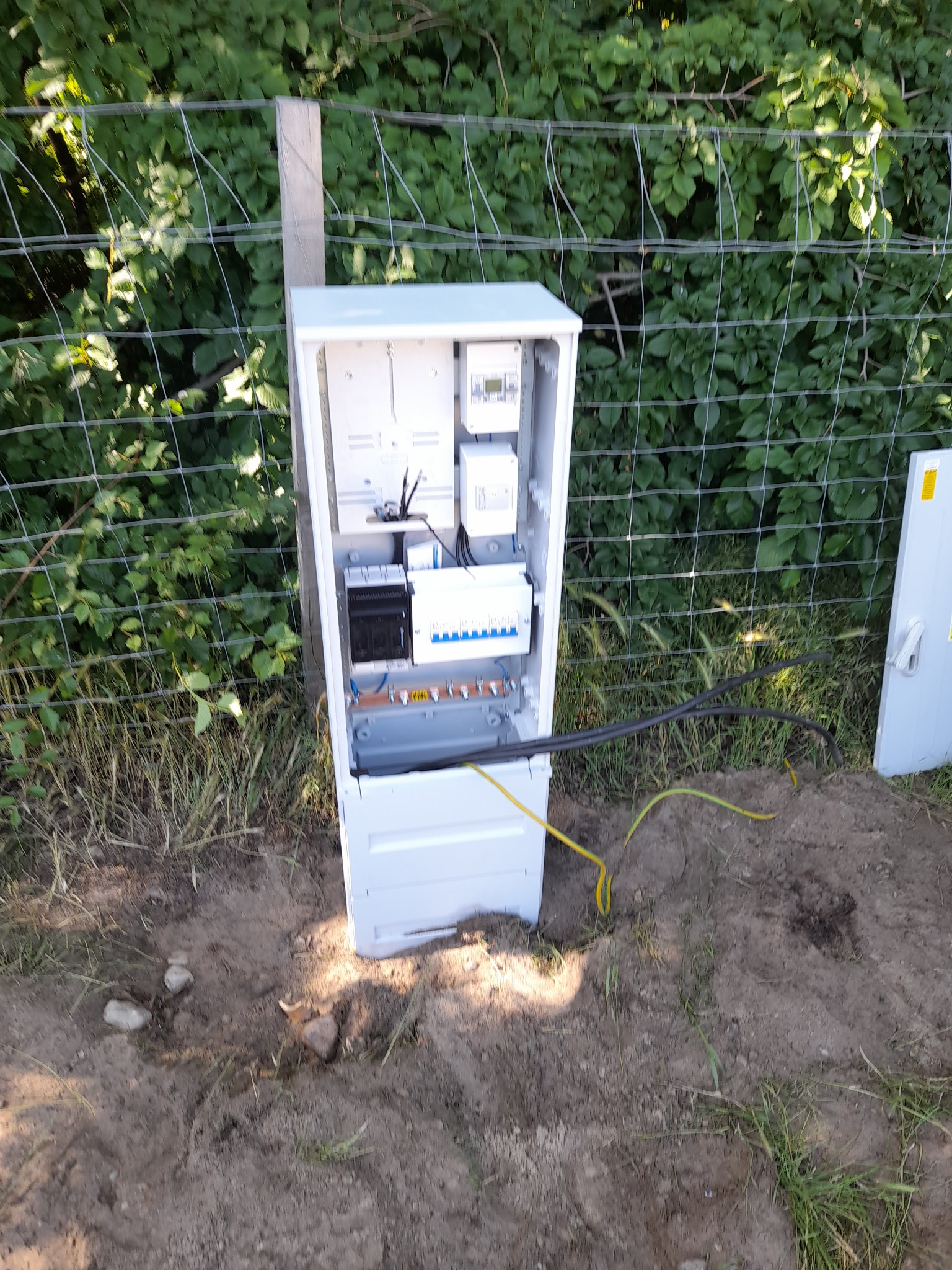 Otwarta, biała skrzynka elektryczna z widocznymi bezpiecznikami i przewodami, umieszczona na ziemi obok ogrodzenia z siatki.
