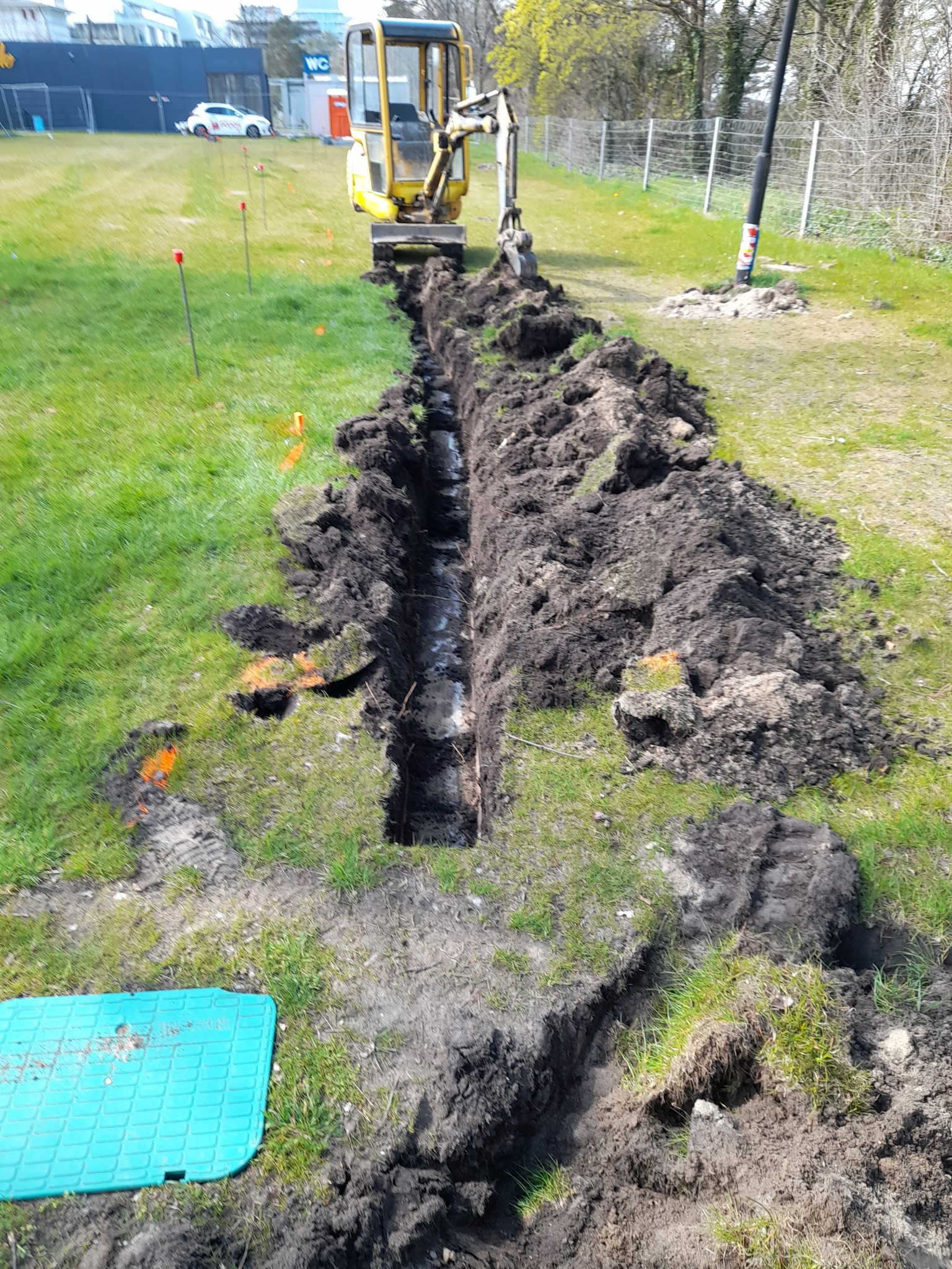 Wykopany rów w trawie, widoczny mały żółty koparka i niebieska pokrywa studzienki. Pomarańczowe znaczniki wzdłuż trasy wykopu sugerują planowaną instalację. W tle budynek i zaparkowany samochód.