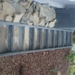 Zewnętrzne schody z szarych płyt, obok skalniak z jasnych kamieni, rabata wysypana korą, widok na ogród i dom w tle.