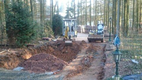 Dwa mini-koparki pracują przy wykopie ziemi w lesie, przygotowując podłoże pod drogę, widoczne lampy oświetleniowe wzdłuż wyznaczonej trasy.