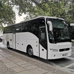 Erfil Trans Wynajem Autokarów, Przewozy Autokarowe, Wynajem Busa, Piaseczno, Warszawa - Nowoczesny, biały autokar turystyczny zaparkowany na brukowanej ulicy, otoczony zielenią drzew. Widoczny fragment chodnika i zaparkowany samochód w tle.