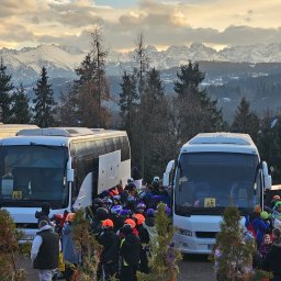 Erfil Trans Wynajem Autokarów, Przewozy Autokarowe, Wynajem Busa, Piaseczno, Warszawa - Autokary z narciarzami na tle górskiego krajobrazu. Grupa osób w zimowych strojach i kaskach stoi przed pojazdami. Widok na ośnieżone szczyty w oddali.