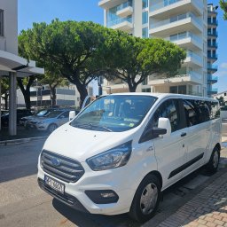 Erfil Trans Wynajem Autokarów, Przewozy Autokarowe, Wynajem Busa, Piaseczno, Warszawa - Biały bus Ford Transit Custom z ciemnymi szybami, zaparkowany na ulicy w słoneczny dzień, z budynkami w tle. Widoczna tablica rejestracyjna i logo firmy.