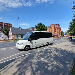 Erfil Trans Wynajem Autokarów, Przewozy Autokarowe, Wynajem Busa, Piaseczno, Warszawa - Biały bus na drodze miejskiej, ceglane budynki w tle, błękitne niebo. Widok z boku, cień drzew na chodniku. Transport osobowy w słoneczny dzień.