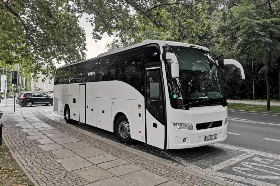 Nowoczesny, biały autokar turystyczny zaparkowany na brukowanej ulicy, otoczony zielenią drzew. Widoczny fragment chodnika i zaparkowany samochód w tle.