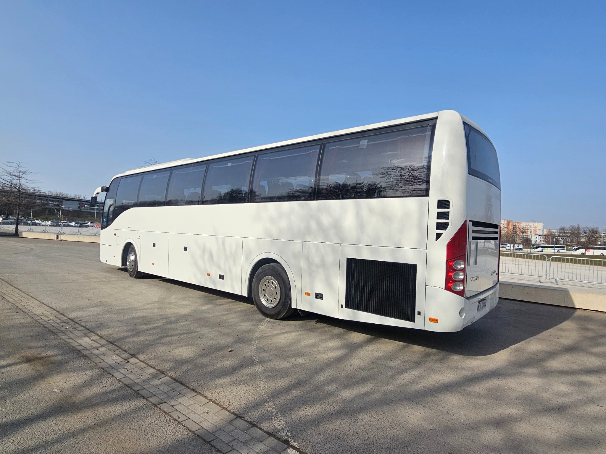 Biały autokar Volvo na parkingu w słoneczny dzień. Widok z tyłu i boku, z widocznymi oknami i elementami karoserii. W tle błękitne niebo i budynki.