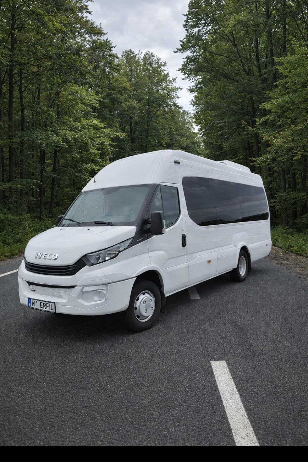 Biały bus Iveco na asfalcie, otoczony zielenią drzew. Pojazd z przyciemnianymi szybami, widoczny z boku, z numerem rejestracyjnym W1 ERFIL. Pochmurne niebo w tle.