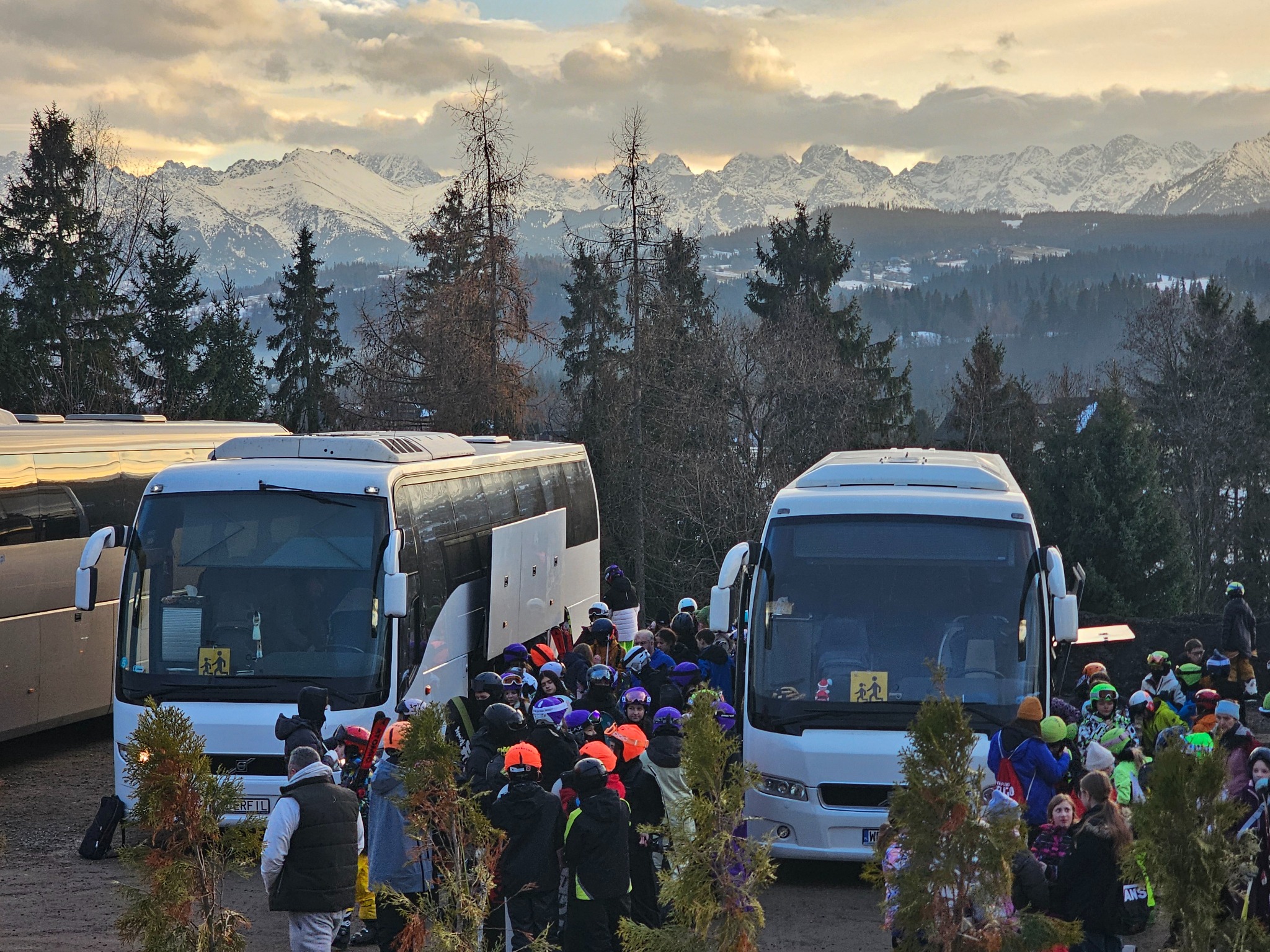 Autokary z narciarzami na tle górskiego krajobrazu. Grupa osób w zimowych strojach i kaskach stoi przed pojazdami. Widok na ośnieżone szczyty w oddali.