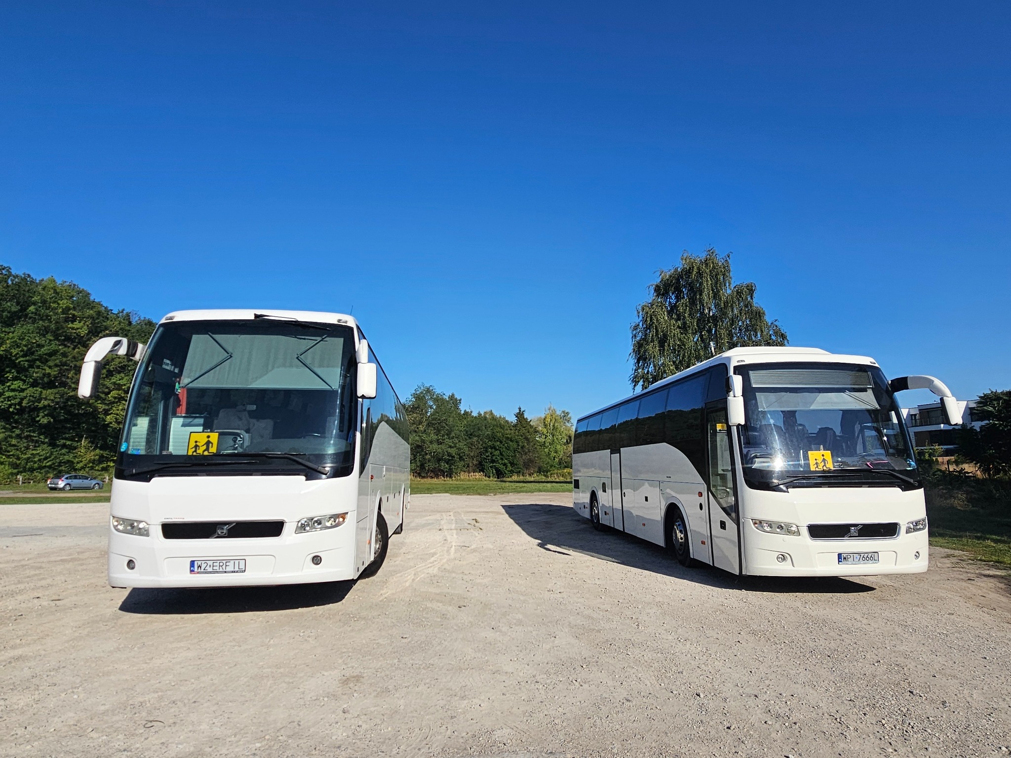 Dwa białe autokary Volvo, jeden z otwartymi drzwiami, stojące na żwirowym parkingu w słoneczny dzień. Tabliczka z symbolem dzieci na przedniej szybie.