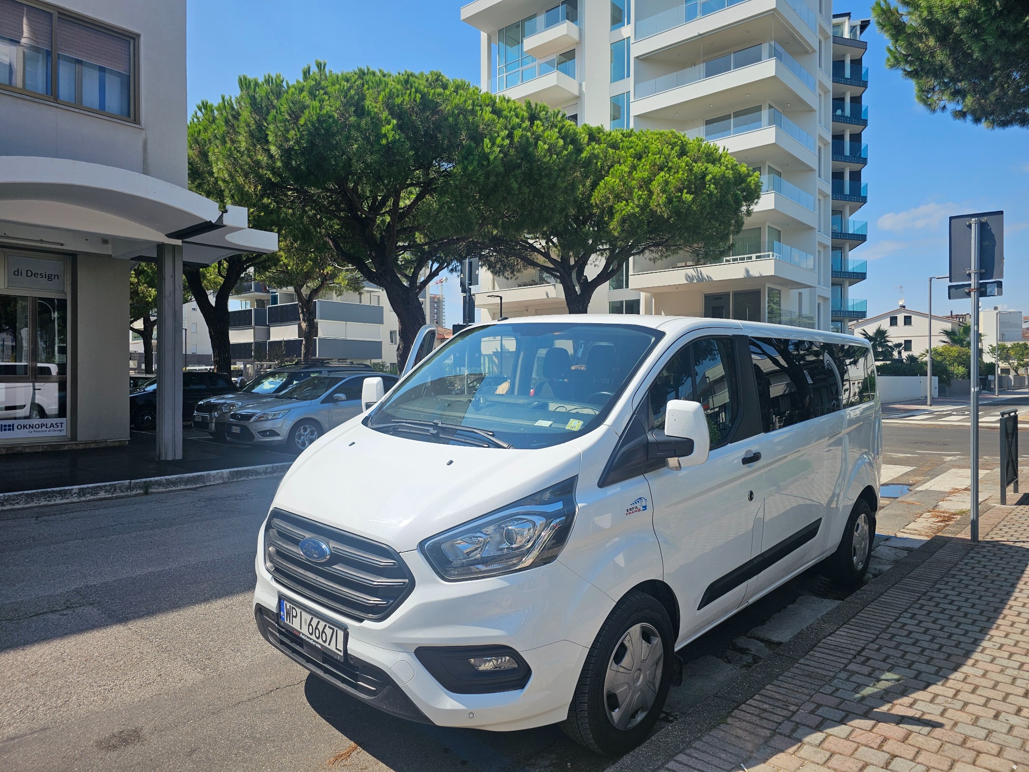 Biały bus Ford Transit Custom z ciemnymi szybami, zaparkowany na ulicy w słoneczny dzień, z budynkami w tle. Widoczna tablica rejestracyjna i logo firmy.