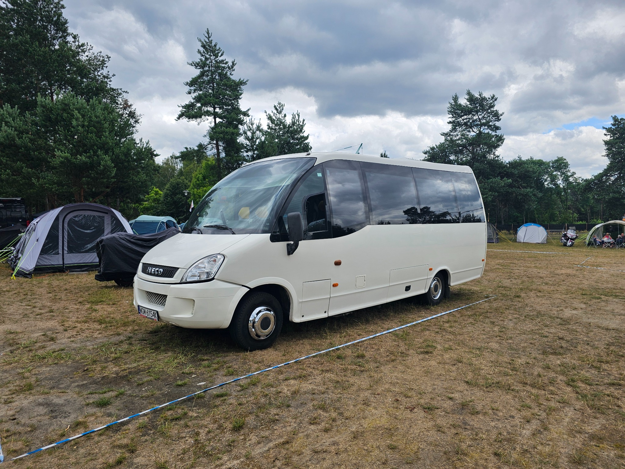 Biały bus Iveco na trawiastym terenie kempingu, w tle namioty i drzewa pod pochmurnym niebem. Widoczny fragment niebiesko-białej taśmy wygradzającej.