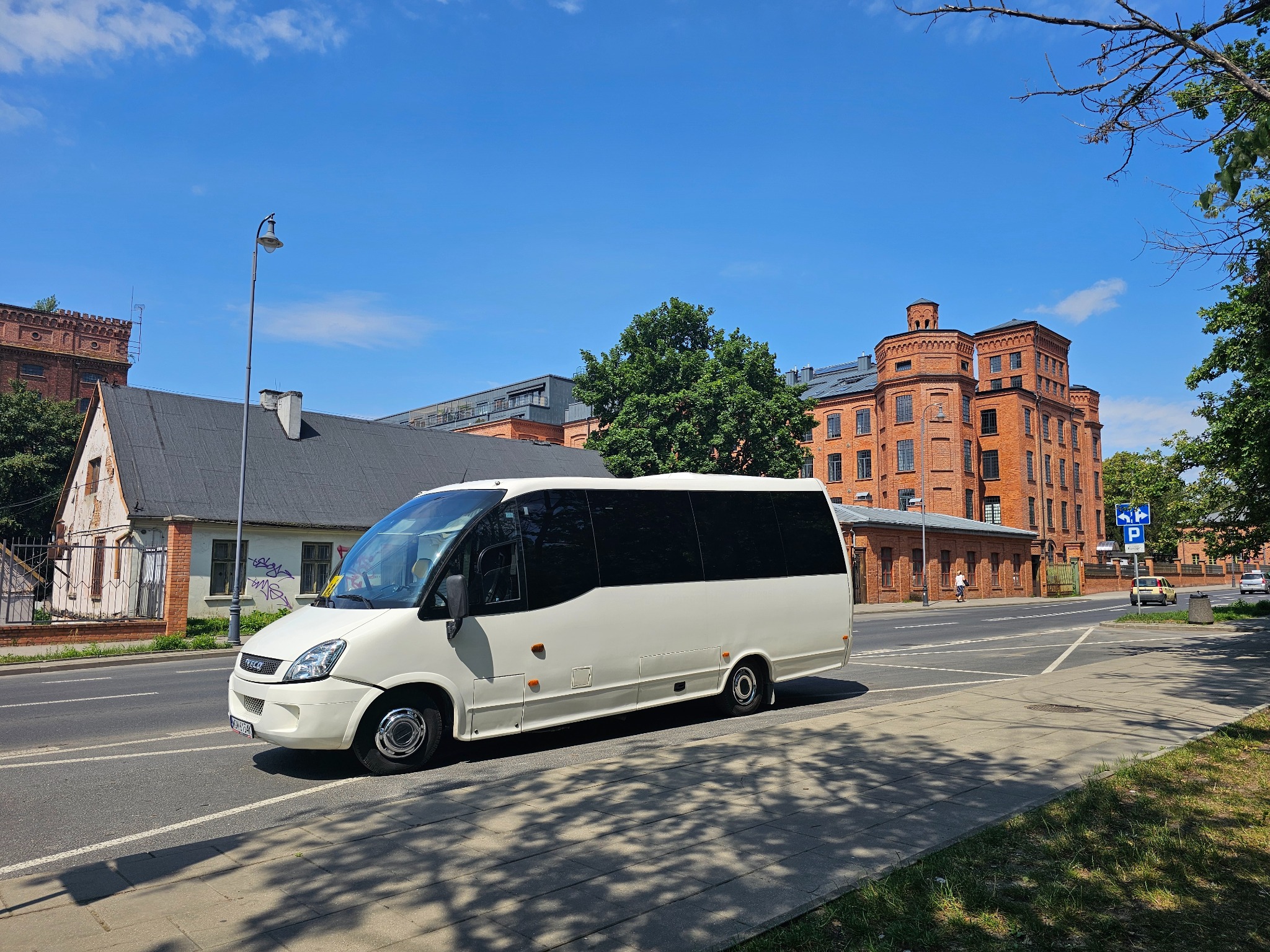 Biały minibus Iveco na tle ceglanych budynków i błękitnego nieba, zaparkowany przy ulicy. Widok z boku, cień drzew na chodniku.