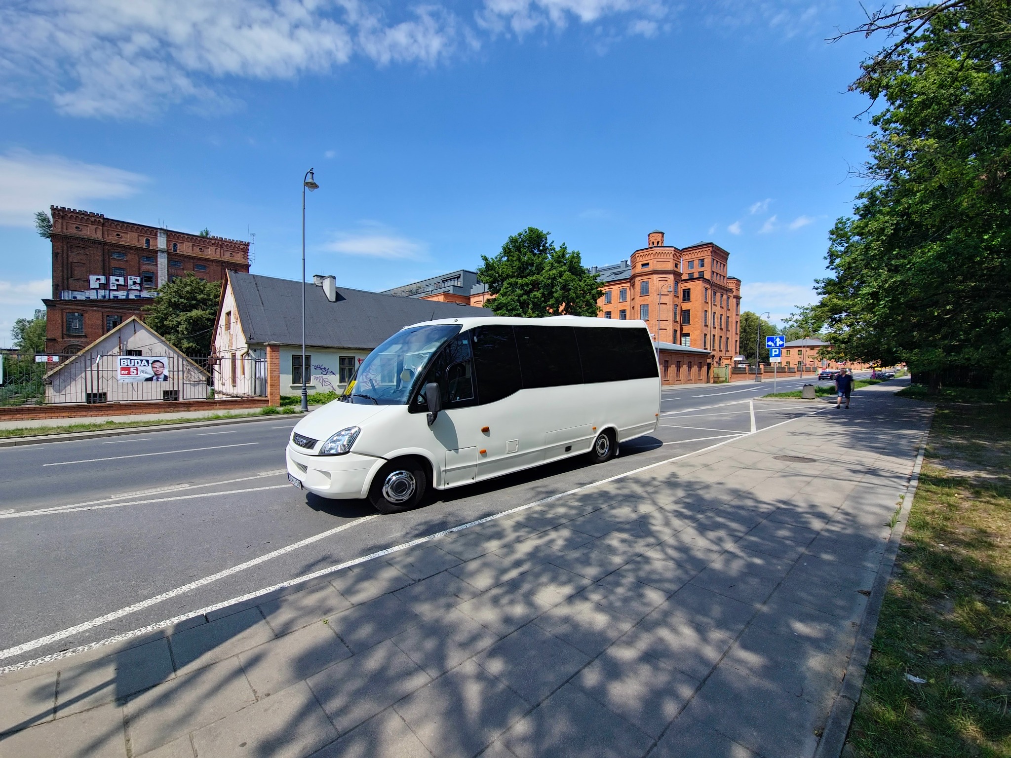 Biały bus na drodze miejskiej, ceglane budynki w tle, błękitne niebo. Widok z boku, cień drzew na chodniku. Transport osobowy w słoneczny dzień.