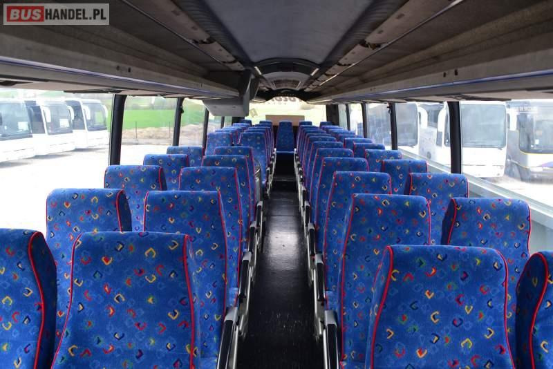 Wnętrze autobusu turystycznego z rzędami niebieskich foteli z kolorowym wzorem, widok z przodu kabiny pasażerskiej, za oknami widoczne inne autobusy.