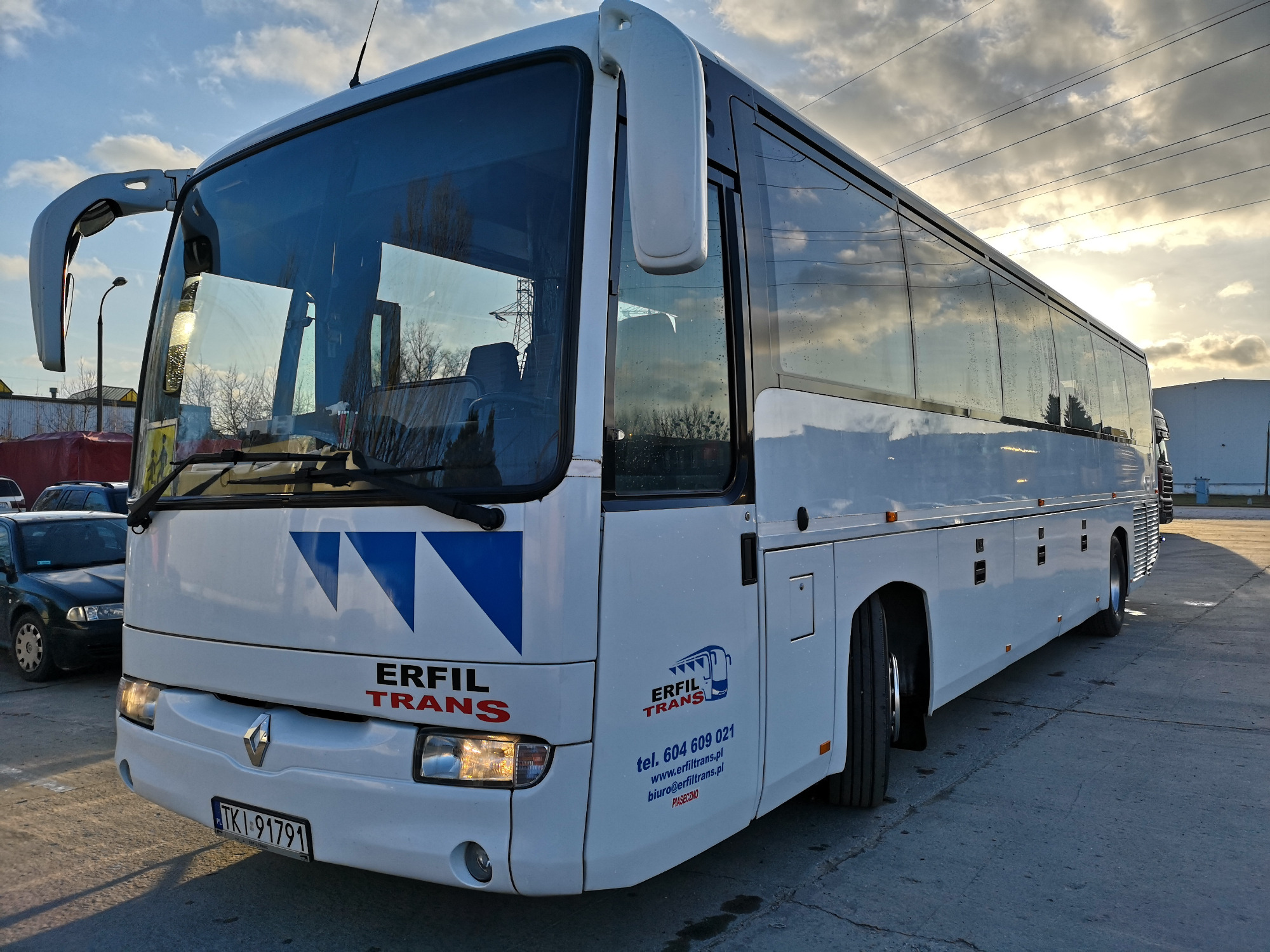 Biały autokar turystyczny marki Renault z niebieskim logo firmy Erfil Trans na parkingu, widoczne tablice rejestracyjne i odbicie chmur w szybach.