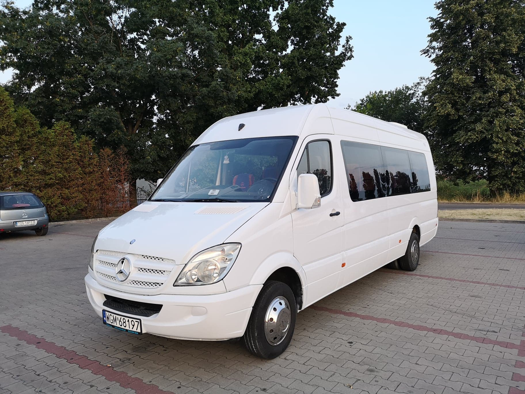Biały Mercedes-Benz Sprinter, widok z przodu i boku, zaparkowany na kostce brukowej z widoczną tablicą rejestracyjną WGM 68197 i chromowanymi kołpakami, drzewa w tle.