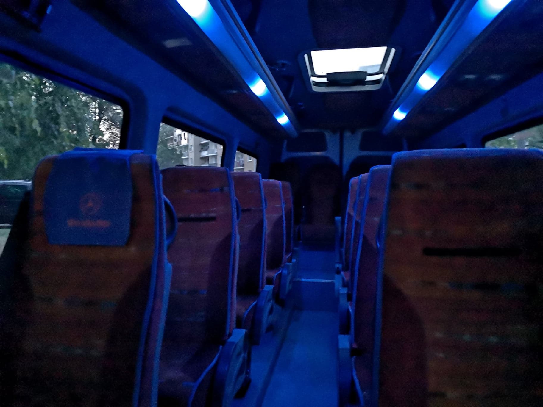 Wnętrze pustego minibusa z rzędami brązowych foteli z niebieskimi zagłówkami z logo Mercedes-Benz, oświetlone niebieskimi lampami LED pod sufitem, widok z tyłu pojazdu.