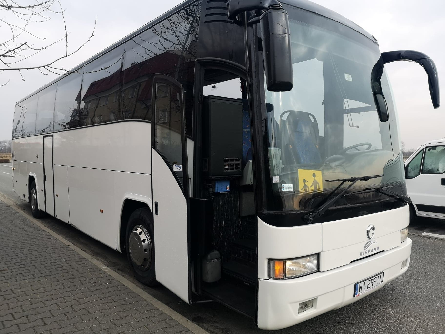 Biały autokar Hispano z otwartymi drzwiami, zaparkowany na chodniku z kostki brukowej, w tle budynek z czerwonym dachem odbijający się w szybach.