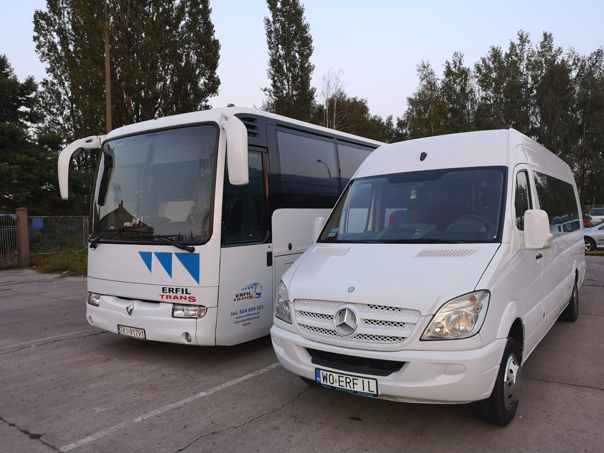 Dwa białe pojazdy: autobus Renault z logo Erfil Trans i biały Mercedes Sprinter, zaparkowane na betonowym placu, z drzewami w tle.