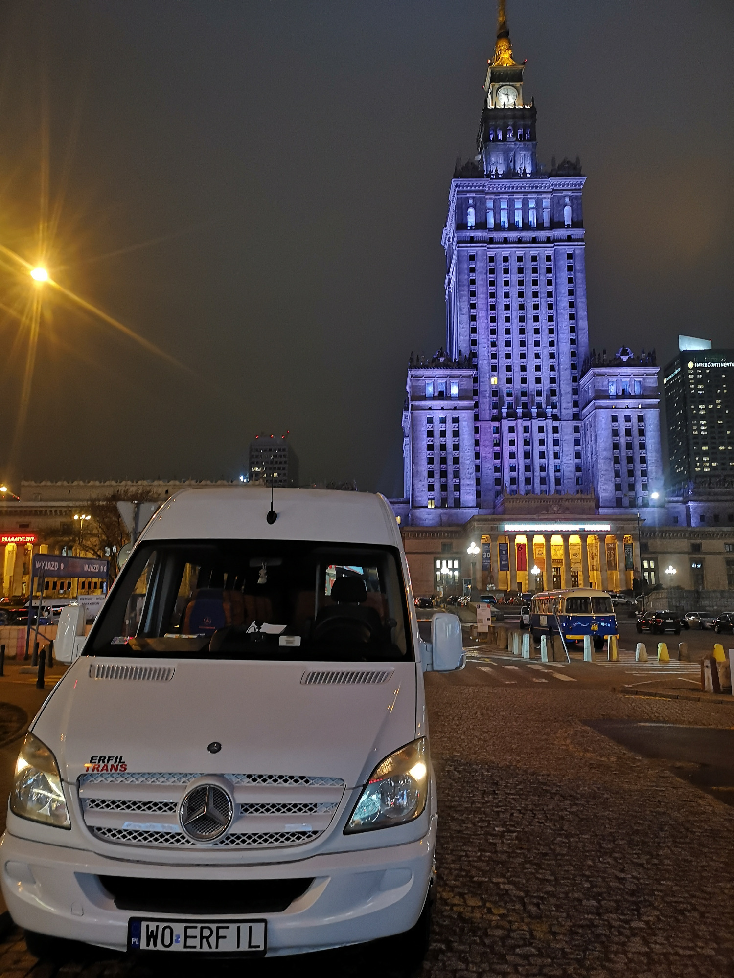 Biały bus Mercedes-Benz z logo firmy Erfil Trans na tle nocnego Pałacu Kultury i Nauki w Warszawie.